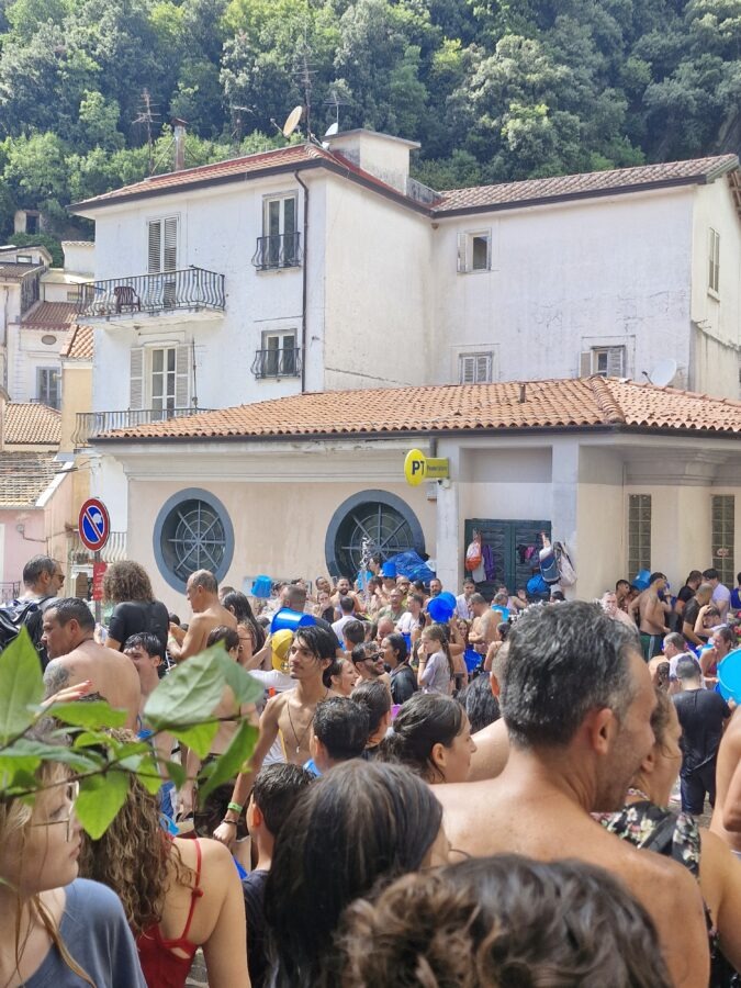 Tłum ludzi biorących udział w wodnej bitwie podczas święta A Chiena w Campagna w Kampanii; uczestnicy z niebieskimi wiadrami oblewają się wodą na tle jasnych budynków i zielonego wzgórza.