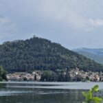 View of a lakeside town at the foot of a forested hill with a castle ruin on the summit, surrounded by mountains.