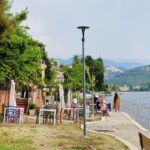 Lakeside promenade with colorful outdoor café chairs, closed umbrellas, and people sitting by the water, with mountains in the background.