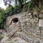 Ancient stone wall with an arched opening, partly built into a rocky hillside, surrounded by greenery and trees.