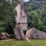 Among the trees and green bushes lie the ruins of a bridge, part of an ancient Roman bridge stands above the green grass