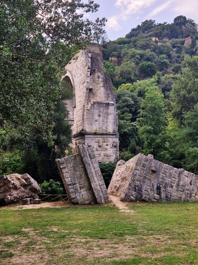 Among the trees and green bushes lie the ruins of a bridge, part of an ancient Roman bridge stands above the green grass
