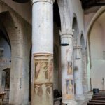 Interior of a Romanesque church with stone columns decorated with medieval frescoes of saints and biblical scenes, wooden benches, and hanging candle chandeliers.