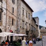 Cobblestone street in an Italian medieval town with old stone buildings, outdoor café tables under umbrellas, and people walking and sitting.