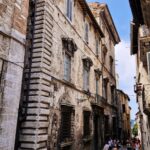Narrow cobblestone street in an Italian medieval town, with tall stone buildings featuring arched windows and iron grilles, and groups of people walking below.