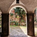 Stone archway passage opening onto a small courtyard with cobblestones, potted plants, ivy-covered walls, and a hanging lantern above.