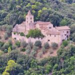 Medieval stone monastery with a bell tower, surrounded by fortified walls and nestled in a dense forest on a hillside.