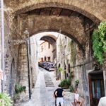 Steep cobblestone alley in a medieval town, framed by large stone archways, with potted plants along the walls and people walking and sitting.