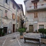 Quiet stone square in an Italian town, surrounded by old houses with wooden shutters, potted plants, and a bench in the center.