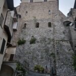 Tall stone building with a fortified base, narrow windows, and potted plants at the foot of the wall, seen from a steep street in an old town.