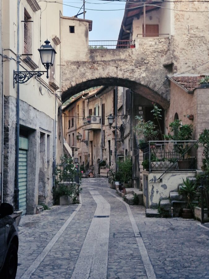 Old stone houses line the cobblestone street on either side. The houses are connected by arches, and pots of green plants and large old lamps hang on the walls.