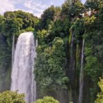 a tall waterfall flows down from a high slope among green plants, and right next to it a narrow stream of water flows down