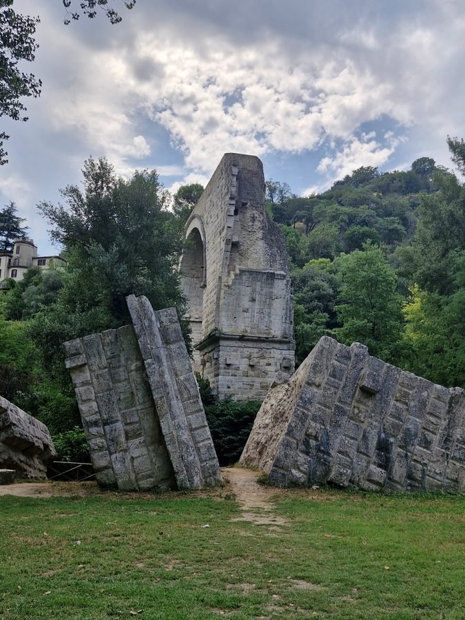Fallen stone blocks of the ancient Roman Ponte d'Augusto on a grassy clearing, with the surviving high arch standing against wooded slopes in Narni, Umbria.