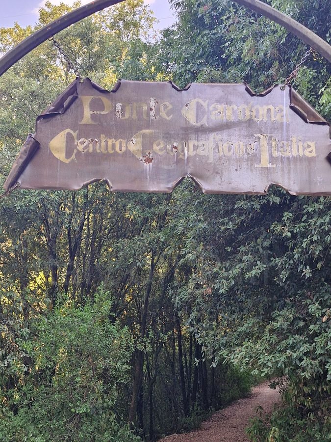 Weathered metal sign reading "Ponte Cardona Centro Geografico d'Italia" hanging over a dirt path through dense woods in Umbria.