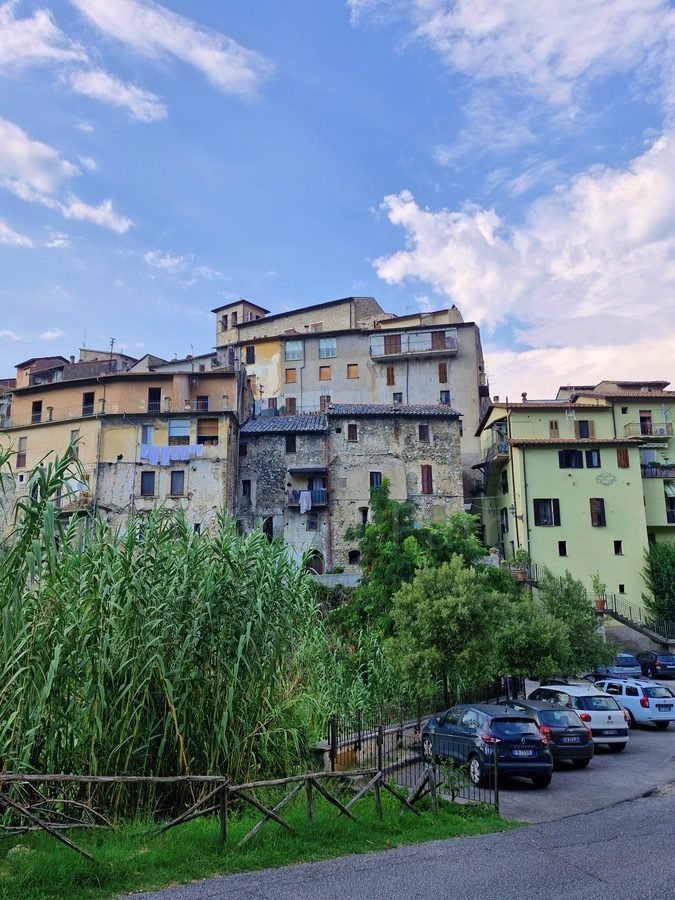 View from a distance of stone and pastel houses in the borgo of Papigno, Umbria, with parked cars and dense vegetation in the foreground.