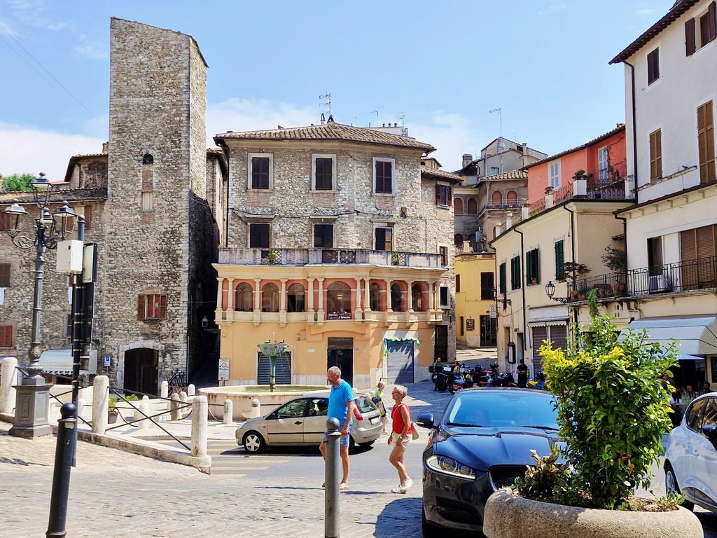 Town square in Narni, Umbria, with a stone medieval tower, central fountain, historic facades, and parked cars under a clear sky.