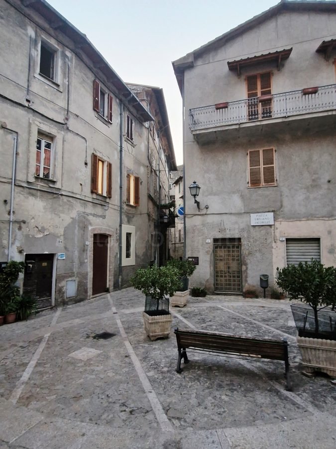 Piazza Emiliano Jacobelli in Papigno, Umbria, with a stone-paved square, wooden bench, potted plants, and weathered residential facades.