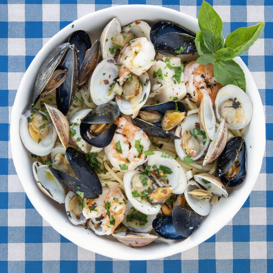 A white bowl filled with spaghetti, mussels, clams, and shrimp, garnished with fresh parsley and basil, placed on a blue and white checkered tablecloth.