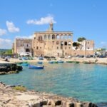 Historic stone building with arched windows and a small bell tower overlooking a turquoise harbor, with colorful fishing boats floating in the water under a bright blue sky.