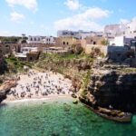 Crowded beach with turquoise water surrounded by cliffs and whitewashed houses in Polignano a Mare, Puglia, Italy.