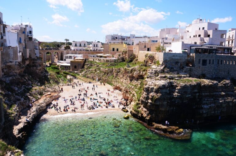 Crowded beach with turquoise water surrounded by cliffs and whitewashed houses in Polignano a Mare, Puglia, Italy.