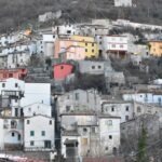 Hillside village in Italy with colorful houses, old stone buildings, and terracotta rooftops stacked along the slope beneath a wooded mountain