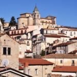 Historic hilltop village in Italy with stone houses, terracotta rooftops, and a clock tower church under a clear blue sky.