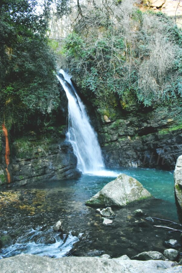 Scenic waterfall in Italy cascading into a turquoise pool, framed by moss-covered rocks and lush greenery, creating a peaceful nature scene
