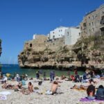 Tourists relaxing on Lama Monachile beach in Polignano a Mare, Puglia, with historic stone houses built on the cliffs above