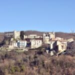 Hilltop village in Italy with a medieval stone castle, church towers, and houses surrounded by winter woodlands under a clear blue sky