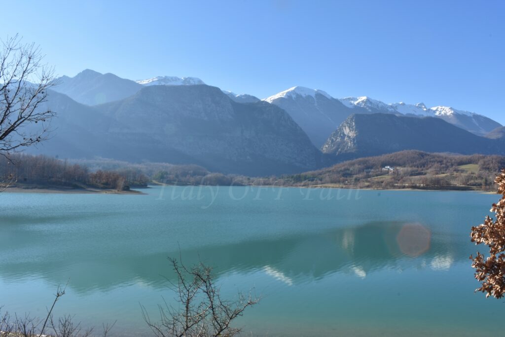 Molise in winter, Clear turquoise lake surrounded by bare winter trees, with snow-capped mountains in the background under a bright blue sky.
