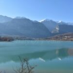 Molise in winter, Clear turquoise lake surrounded by bare winter trees, with snow-capped mountains in the background under a bright blue sky.