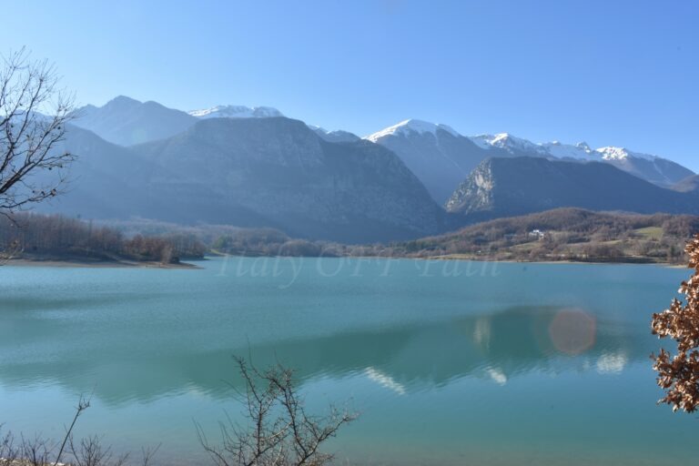 Molise in winter, Clear turquoise lake surrounded by bare winter trees, with snow-capped mountains in the background under a bright blue sky.