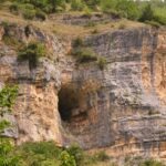 The photo shows a hiker pausing on a narrow mountain trail in Majella National Park. She leans against a large rock, wearing a sun hat, sunglasses, and hiking gear with trekking poles and a camera around her neck. Behind her, the landscape opens into a sweeping view of green valleys and rugged hillsides, dotted with patches of yellow wildflowers. The trail is rocky but framed by lush vegetation, giving a sense of both challenge and reward. The image captures the spirit of exploration and the beauty of hiking in the wild Italian mountains
