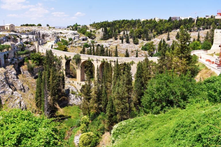 Ancient stone aqueduct surrounded by cypress trees in Gravina di Puglia, Italy, with rocky cliffs and historic buildings in the background in Gravina in Puglia