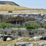 Ancient rock-cut caves and columns carved into the cliffs of Gravina di Puglia, Italy, with olive trees and stone walls in the foreground