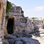 Ancient cave dwelling carved into limestone rock in Gravina di Puglia, Italy, with a dark entrance surrounded by rugged stone walls