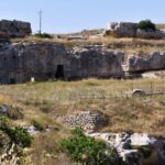 Rock-cut caves and ancient dwellings in the limestone cliffs of Gravina di Puglia, Italy, surrounded by wild grass and stone walls.