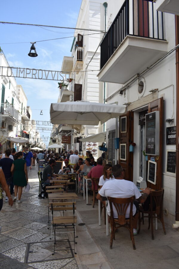 A lively Italian street lined with white buildings, outdoor cafés, and people dining under umbrellas, with pedestrians walking along the cobblestone path on a sunny day.