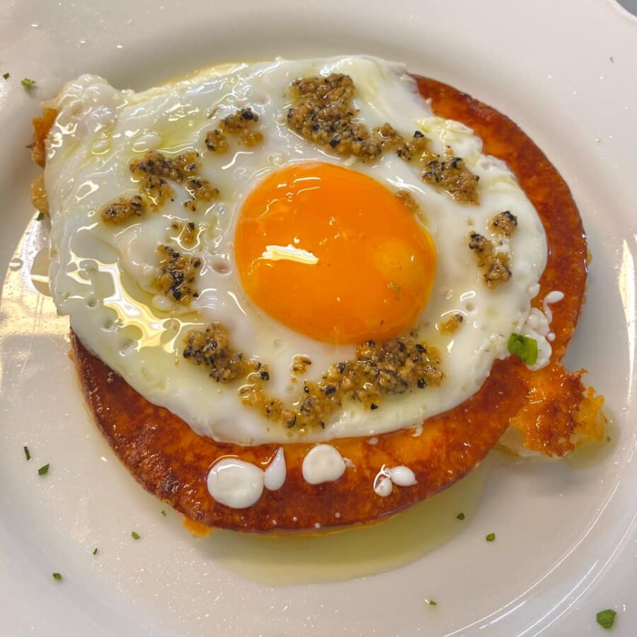 Where to eat in Castelpetroso Fried egg with a bright orange yolk served on top of a crispy round slice of fried cheese, garnished with minced garlic and herbs.