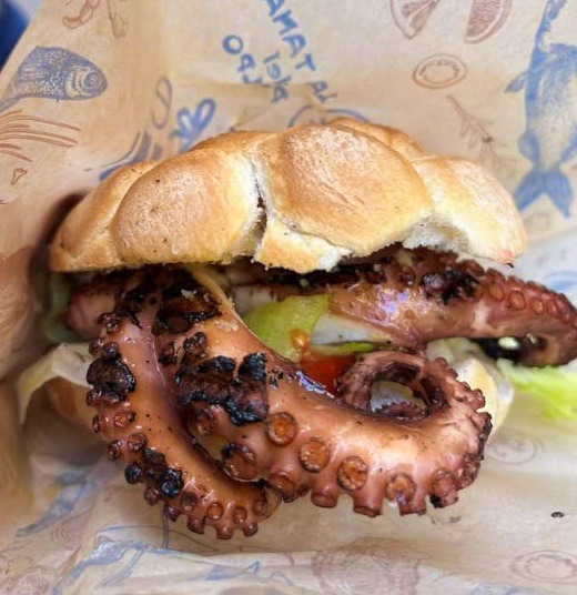 A sandwich filled with grilled octopus tentacles, lettuce, and tomato, served in a crusty bread roll and wrapped in paper.
