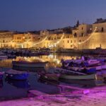 Evening view of Bisceglie’s harbour with small fishing boats and warm string lights reflecting on the water during Calici nel Borgo Antico.