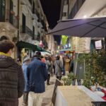 Nighttime crowd walking through the narrow streets of Bisceglie’s old town during Calici nel Borgo Antico, with stalls, decorations and grape-shaped balloons overhead.