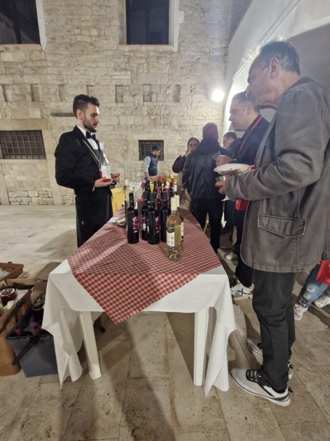 A sommelier serving wine at a tasting table during Calici nel Borgo Antico in Bisceglie, with visitors sampling different bottles in the old town.