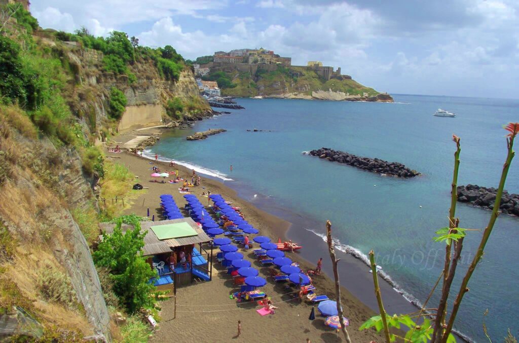 View of Pozzo Vecchio Beach on Procida Island, Italy, with rows of blue umbrellas, golden sand, and the historic Terra Murata hill in the background.