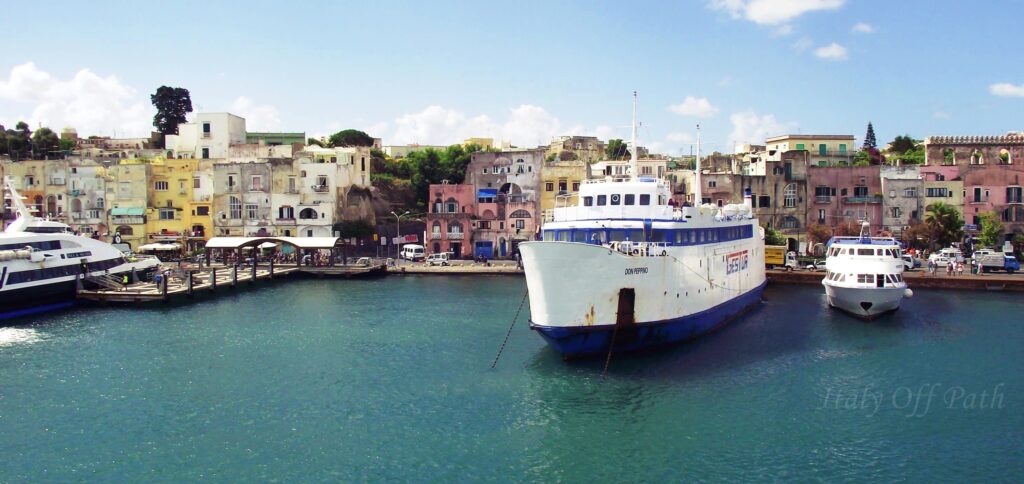 Colorful waterfront buildings and lively street scene at the Marina Grande port on Procida Island, Italy, with cars, scooters, and people in front of pastel houses.