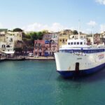 Colorful waterfront buildings and lively street scene at the Marina Grande port on Procida Island, Italy, with cars, scooters, and people in front of pastel houses.