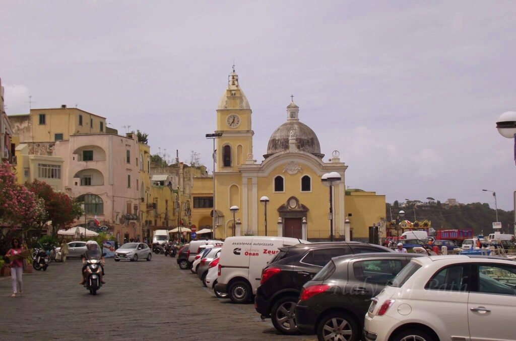 The yellow Church of Santa Maria della Pietà at Marina Grande port on Procida Island, Italy, surrounded by pastel buildings, cars, and scooters.