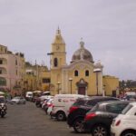 The yellow Church of Santa Maria della Pietà at Marina Grande port on Procida Island, Italy, surrounded by pastel buildings, cars, and scooters.