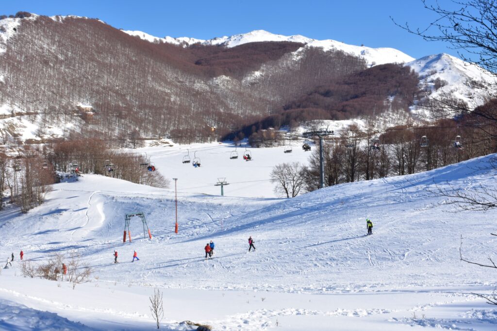 Skiers on a sunny winter slope in Campitello Matese, with snow-covered mountains and chairlifts in the background.
